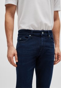Jean en denim bleu foncé coupe slim ; comprend une fermeture par bouton et fermeture éclair, deux poches avant, et un logo de marque discret sur la poche.