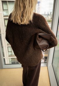 Blonde vrouw staat bij een raam, draagt een pluizige bruine trui en broek, houdt een bruine leren tas onder haar arm.