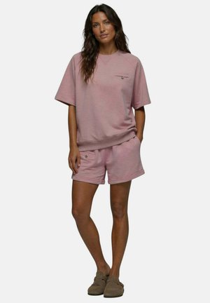 Femme aux longs cheveux bruns portant un haut rose à manches courtes ample et un short assorti avec des poches, debout une main dans la poche et des chaussures marron.