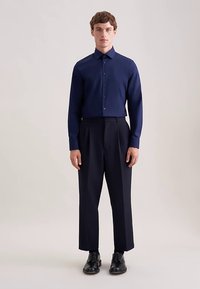 Camicia blu navy a maniche lunghe con collo button-down, abbinata a pantaloni scuri a pieghe e scarpe nere stringate. Tessuto dalla texture liscia.