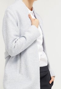 Giacca azzurro chiaro, con texture, colletto a scialle, maniche lunghe e tasche con zip. Indossata sopra una camicia bianca e pantaloni scuri.