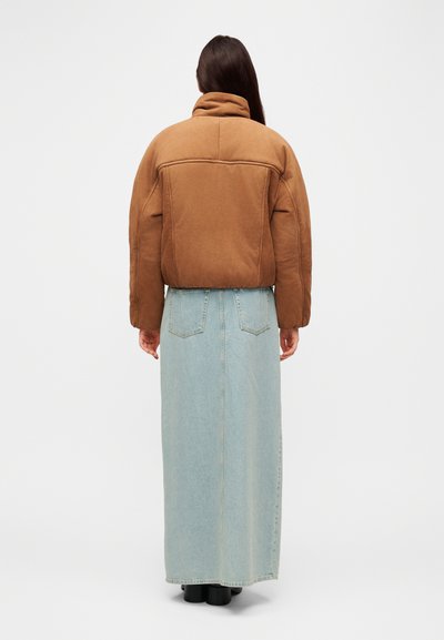 Veste matelassée marron avec col haut, associée à une jupe longue en denim bleu clair. Vue de dos montrant des coutures ajustées et un équipement minimal.