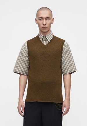 Homme portant un pull sans manches marron par-dessus une chemise à manches courtes à carreaux et un pantalon noir, debout sur fond blanc.