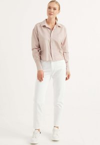 Camicia button-up cropped rosa chiaro con colletto, abbinata a jeans slim-fit bianchi e sneakers bianche. Tessuto morbido con maniche lunghe.