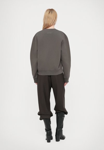Persona con cabello largo y rubio de espaldas, vistiendo una sudadera gris oscuro, pantalones oscuros holgados y botas negras de tacón alto hasta la rodilla.