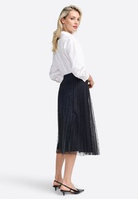 Jupe plissée noire en tulle avec un motif à pois, associée à une chemise blanche à boutons. Des escarpins noirs à talons ouverts complètent la tenue.