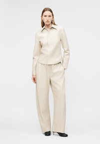 Žena oblečená v béžovej koženej košeli s dlhými rukávmi a širokými nohavicami, stojaca s jednou rukou v vrecku, čierne lodičky na bielom pozadí.