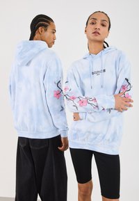 Hoodie em tie-dye azul claro com bordados florais nas mangas, apresentando um capuz com cordão e bolso canguru. Calções pretos usados com o hoodie.