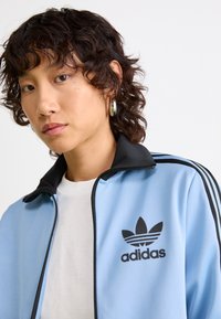 Jaunas žmogus su trumpais garbanotais plaukais, apsirengęs šviesiai mėlynu Adidas treniruočių džemperiu su juodomis juostelėmis ir baltu marškinėliu, fone esant paprastai. 