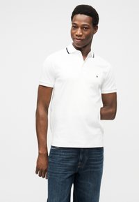 Λευκό κοντομάνικο πόλο με μαύρο περιθώριο στο γιακά, φορεμένο με μπλε τζιν. Υπάρχει διακριτικό λογότυπο στο στήθος. Ομαλή υφή υφάσματος.