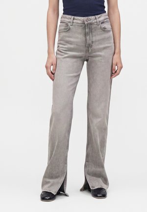 Femme portant un jean gris clair taille haute avec des fentes à l'avant de l'ourlet et des chaussures noires pointues, assorti à un haut foncé.