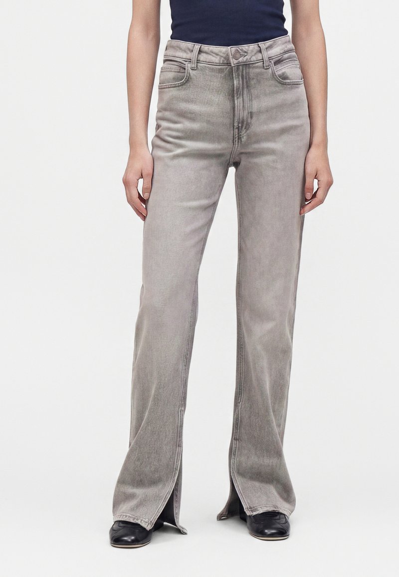 Femme portant un jean gris clair taille haute avec des fentes à l'avant de l'ourlet et des chaussures noires pointues, assorti à un haut foncé.