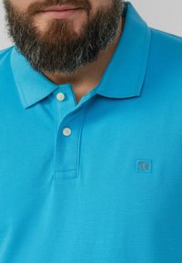 Polo turquoise en tissu texturé, avec une patte de boutonnage à trois boutons, un col plat et un petit logo dans le coin supérieur gauche.