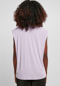 Světle fialový bezrukávový top s volným střihem, měkký bavlněný materiál a zaoblený lem. Zadní část má hladké švy a minimální detaily.