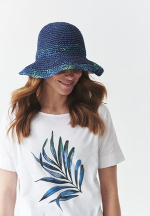 Cappello da sole all'uncinetto blu navy con una larga tesa, caratterizzato da dettagli verdi e blu. Indossato con una camicia bianca che presenta un design di foglia di palma blu.