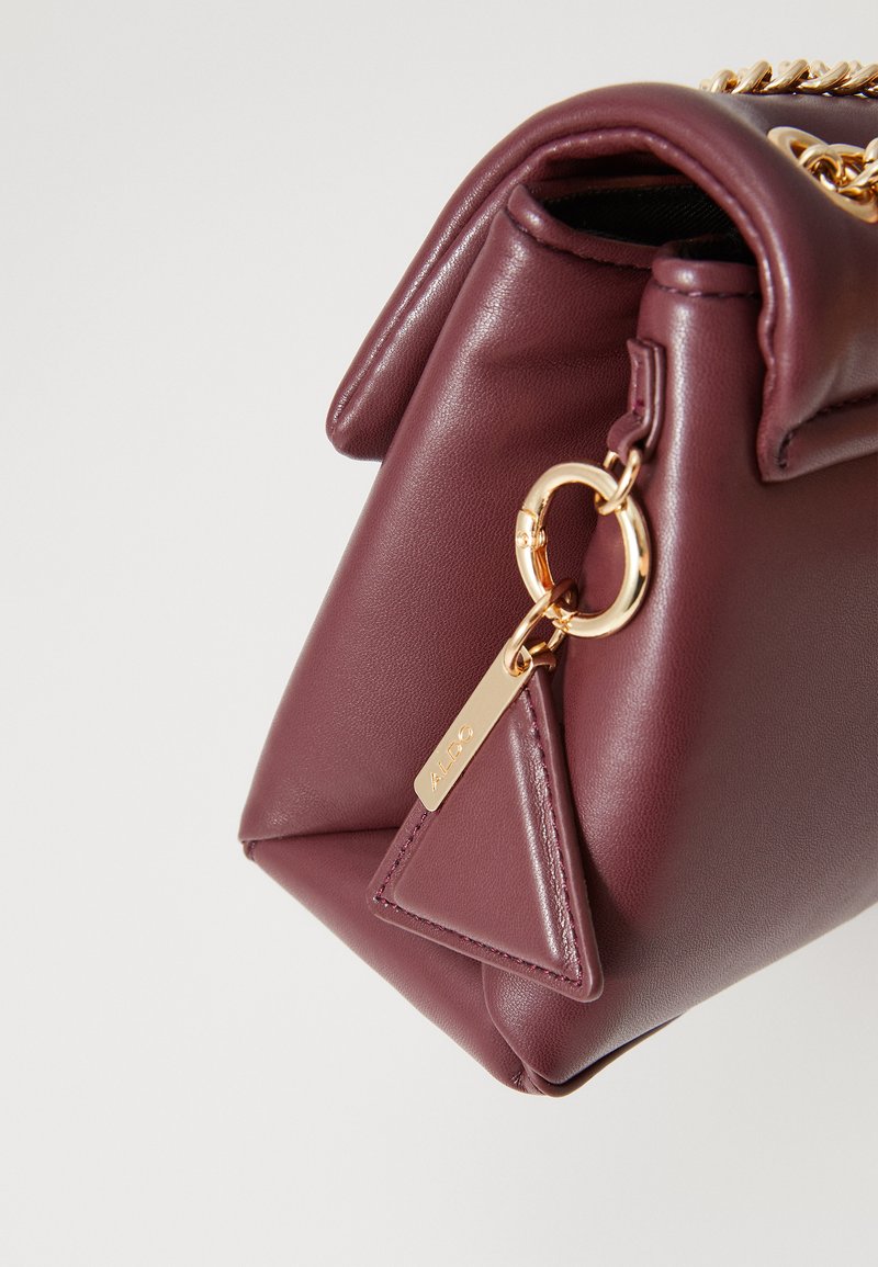 Bordeaux leren handtas met goudkleurige hardware, kettinghengsel en een driehoekige hanger met een logo-tag. Soepele textuur met een gestructureerde vorm.