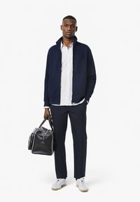 Marine blauw zip-up jas over een wit gestreept shirt, gecombineerd met marineblauwe broek en witte sneakers. Een zwarte tas met witte accenten wordt in de hand gehouden.