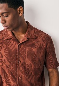 Chemise en éponge à manches courtes couleur rouille avec motifs tactiles texturés. Comprend un col, une patte de boutonnage devant et une poche poitrine.