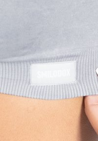 Grijze geribbelde stoffen tanktop met een wit label met "SMILODOX" op de zoom. Toont textuur en stikseldetails.
