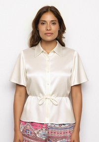 Zijde blouse in crème met een kraag, korte mouwen, knopen aan de voorkant en een koord aan de taille; gestyled met kleurrijke, gedecoreerde broek.