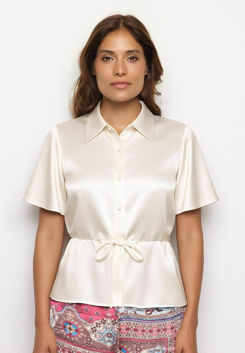 Zijde blouse in crème met een kraag, korte mouwen, knopen aan de voorkant en een koord aan de taille; gestyled met kleurrijke, gedecoreerde broek.