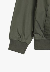 Zielona rękaw kurtki z ribbowanym mankietem i małą czarną metką z napisem "ELEMENT" blisko szwu kieszeni.