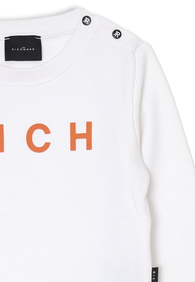 Felpa bianca con il testo arancione "RICH". Presenta due bottoni neri con il logo "JR" sulla spalla. Tessuto morbido ed elastico con scollatura a costine.
