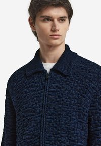 Marinblå stickad jacka med ett texturerat mönster, dragkedja fram och bred krage. Tyget verkar mjukt och tjockt, vilket gör det lämpligt för kallare väder.