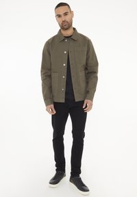 Oliven grøn jakke med knaplukning, to forlommer og en brystlomme, parret med sorte jeans og sorte sneakers.