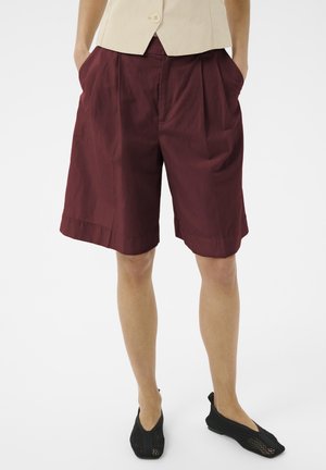 Persona che indossa pantaloncini color granata fino al ginocchio, top beige e scarpe piatte nere intrecciate, in piedi con le mani nelle tasche su sfondo bianco.