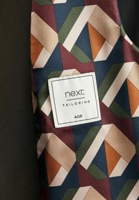Eine maßgeschneiderte Weste mit geometrischen Mustern in Rot, Grün und Braun, mit einem an der Seite genähten Etikett, auf dem "next TAILORING 40R" steht.