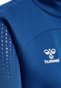 Blå stof sports-top med teksturerede ærmer, der har cirkulære mønstre, og et hvidt logo med et bi-lignende design på brystet.
