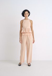 Conjunto de cor pêssego com uma blusa sem mangas com camadas de babados e calças largas, confeccionado em um tecido suave. Sapatilhas pretas completam o visual.