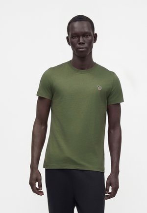 Mann trägt ein olivgrünes kurzärmeliges T-Shirt mit einem kleinen mehrfarbigen Zebralogo auf der linken Brust und steht vor einem einfarbigen weißen Hintergrund.