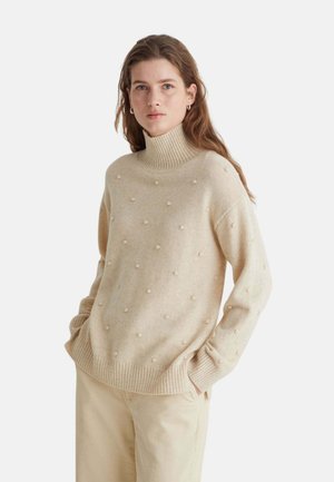 Mujer con cabello largo que lleva un suéter de cuello alto beige con pequeños puntos texturizados y pantalones beige claro, de pie con las manos en los bolsillos.