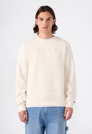 Junger Mann mit mittel-langem dunklem Haar, der einen cremefarbenen Champion-Sweatshirt und hellblaue Jeans vor einem schlichten weißen Hintergrund trägt.