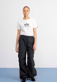 Valkoinen t-paita, jossa on musta "Alpha Industries" -logo, yhdistettynä löysiin mustiin housuihin. Pehmeä kangas, jossa on joustava vyötärö. Yksinkertainen muotoilu.