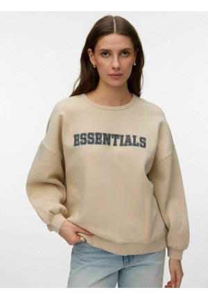Jeune femme aux longs cheveux bruns portant un sweat-shirt beige avec le texte "ESSENTIALS" et un jean bleu clair, debout devant un fond blanc.