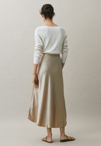 Vrouw met haar in een bun, gekleed in een witte langslavige top, een beige zijden midi-rok en tan flatsandalen, staand met haar gezicht naar een effen achtergrond.