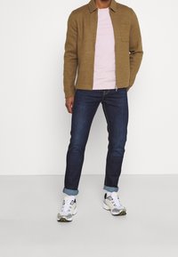 Brun ullzipjacka, ljusrosa t-shirt, indigoblå jeans, uppvikta ärmar, och vita/grå sneakers med gula detaljer.