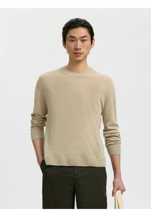 Jonge man met een beige trui en donkere broek staat voor een effen achtergrond en houdt een lichtgekleurde draagtas in zijn rechterhand.
