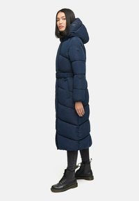 Lång marinblå puffjacka med huva, bälte i midjan och chevron-quiltat mönster. Bärs med svarta jeans och snörade kängor.