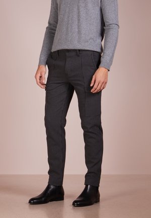 Mand iført tætsiddende mørkegrå ternede bukser, grå langærmet sweater og sorte læderankelstøvler, der står på en beige gulv.