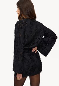 Zwarte textuur jurk met lange klokmouwen, voorzien van een doorschijnende overlay en gegraveerde patronen, strak om de taille.