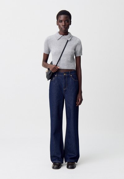 Suéter gris de manga corta con cuello de perlas, combinado con jeans de mezclilla oscuros de pierna ancha y un bolso negro de hombre. Unos zapatos negros sencillos completan el look.