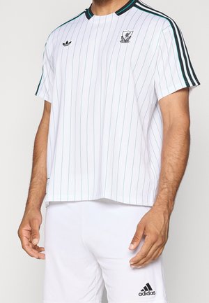 Homme portant une tenue de sport blanche Adidas avec des fines rayures vertes, des logos Adidas et le blason du club de football de Liverpool sur le maillot.