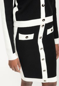 Schwarzes und weißes gestricktes Kleid mit körperbetontem Schnitt, dekorativen Knöpfen und kontrastierenden Taschendetails. Texturierter Stoff mit gerippten Details.