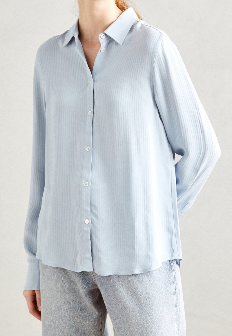 Camicia azzurra chiaro con bottoni in un morbido tessuto a coste, dotata di colletto classico e maniche lunghe, abbinata a jeans in denim chiaro.