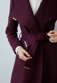 Cappotto in lana bordeaux con collo a scialle, vita con cintura e dettagli in metallo color oro. Presenta una texture liscia e una vestibilità sartoriale.