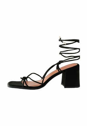 Sandale à talons hauts en daim noir avec un bout carré, des brides croisées et un large talon bloc. Présente des détails de lacets enveloppants.